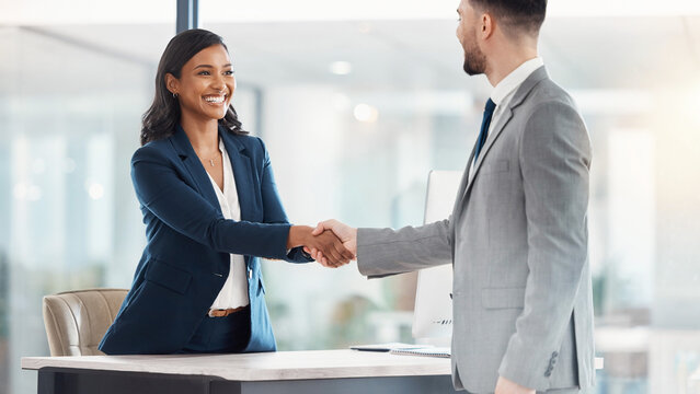 Female HR manager shaking hands with candidate on desk with blurred corporate office background banner.