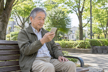 Obraz premium 近所の公園のベンチでスマートフォンを使う日本人シニア男性