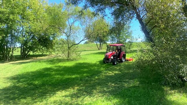 Red agricultural tractor equipped with a mower cuts grass in a vibrant green field, with trees providing shade and a clear blue sky visible in the background. Slow motion footage