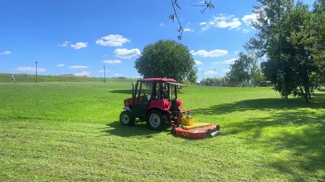 Red agricultural tractor equipped with a mower cuts grass in a vibrant green field, with trees providing shade and a clear blue sky visible in the background. Slow motion footage