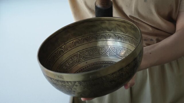 Close up of a woman striking a Tibetan singing bowl and running a mallet along its rim to sustain tones. The etched surface and hand motion are in focus while the background is softly blurred Natural 