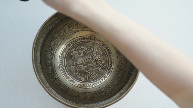 Close up of a hand striking a Tibetan singing bowl filled with water and running a mallet along the rim. The impact and rim motion create visible ripples and vibrating patterns on the liquid surface. 