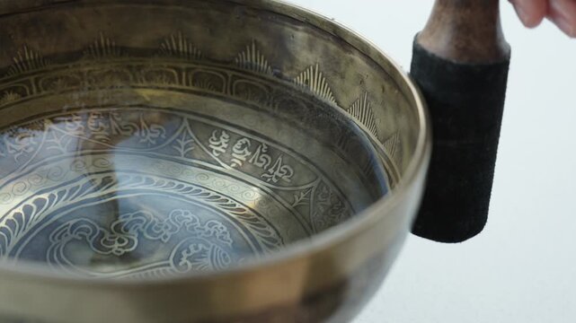 Close up of a hand striking a Tibetan singing bowl filled with water and running a mallet along the rim. The impact and rim motion create visible ripples and vibrating patterns on the liquid surface. 
