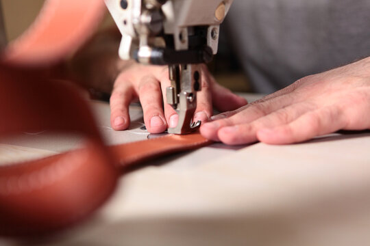 Close-up hands guide red leather under industrial needle. Fingers control feed. Presser foot holds hide. Leathercraft workshop