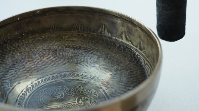 Close up of a hand striking a Tibetan singing bowl filled with water and running a mallet along the rim. The impact and rim motion create visible ripples and vibrating patterns on the liquid surface. 
