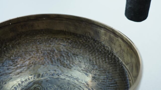 Close up of a hand striking a Tibetan singing bowl filled with water and running a mallet along the rim. The impact and rim motion create visible ripples and vibrating patterns on the liquid surface. 