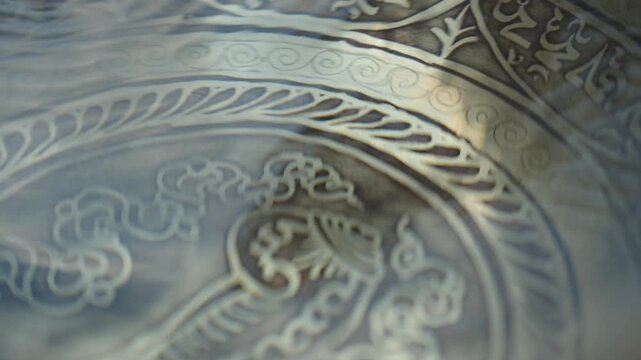 Close up of a hand striking a Tibetan singing bowl filled with water and running a mallet along the rim. The impact and rim motion create visible ripples and vibrating patterns on the liquid surface. 
