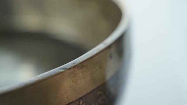 Close up of a hand striking a Tibetan singing bowl filled with water and running a mallet along the rim. The impact and rim motion create visible ripples and vibrating patterns on the liquid surface. 