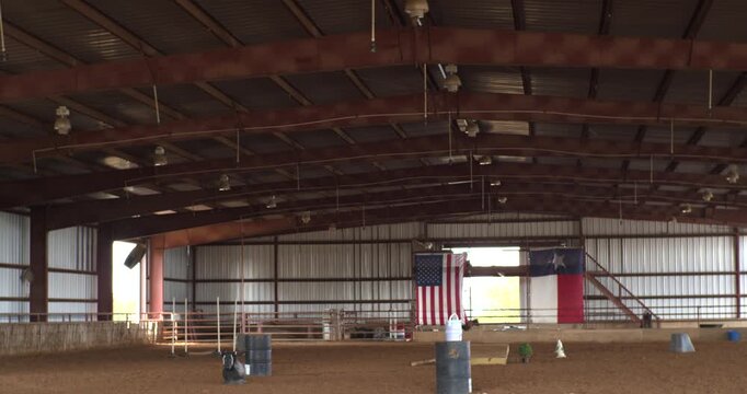 Wide view inside a spacious Texas equestrian arena with metal beams, dirt flooring, ranch equipment, and American and state flags, suitable for western venue themes.