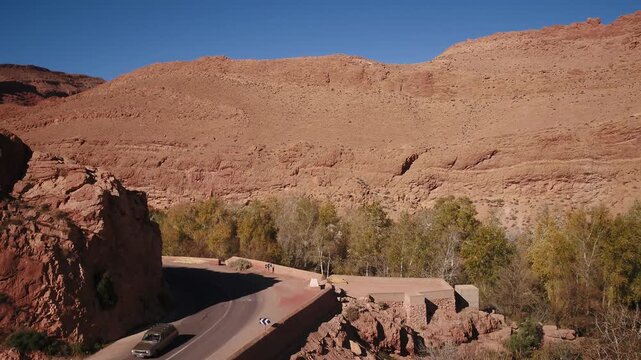 Cinematic aerial view following a paved curved road and bridge carved through rugged canyon rocks and trees in Todra Valley near Tinghir, Morocco.