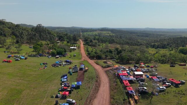 Drone shot of a vibrant camping rally along a dirt road in Nueva Alborada, Paraguay. Colorful tents and cars gather in a lush green rural landscape.