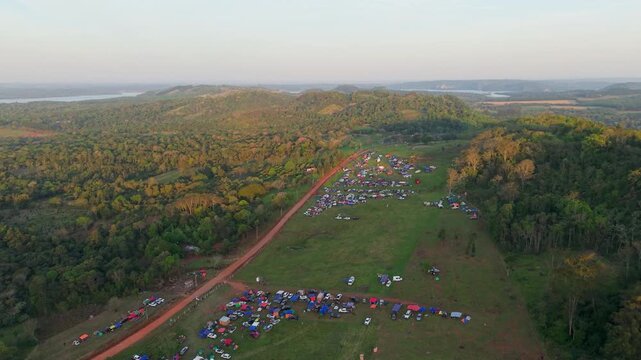 High-altitude drone shot of a sprawling camping rally during golden hour in Nueva Alborada, Paraguay. Hundreds of tents dot a vast green landscape
