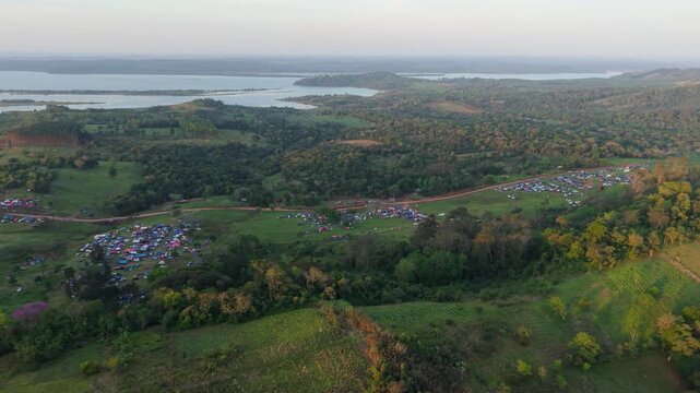 Drone shot of a massive camping rally in Nueva Alborada, Paraguay. Thousands of tents gather in lush green hills with a large river in the background