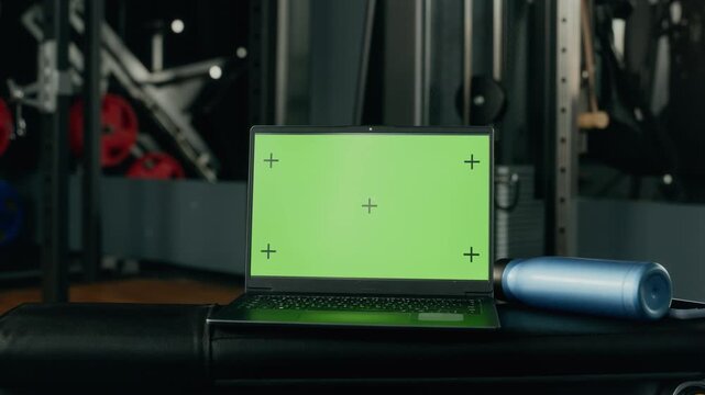 Open laptop with green chroma key screen mockup resting on bench in modern gym. Clip useful for digital fitness software advertising, online workout platform, and athletic app presentation