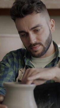 Zoom-in of skilled young man wearing dirty apron making vase from clay in pottery workshop. Worker concentrated on creative activity.