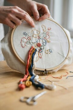 Hands working on a wooden embroidery hoop with colorful thread stitching on linen fabric with sewing scissors and needles on a table