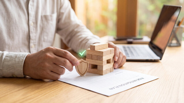 Risk mitigation plan and business protection concept with man placing wooden shield beside block tower on document at laptop desk for insurance strategy, crisis control and safe planning