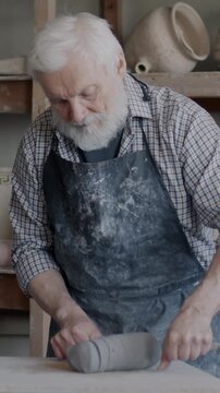 Skilled senior potter working with clay preparing to create ceramic goods in workshop. Elderly man in dirty apron cutting loam with wire then mixing material.