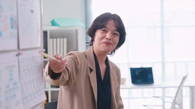 Asian businesswoman sitting on a chair pointing at a presentation board, explaining data and strategy to colleagues during a team meeting in a modern corporate office.