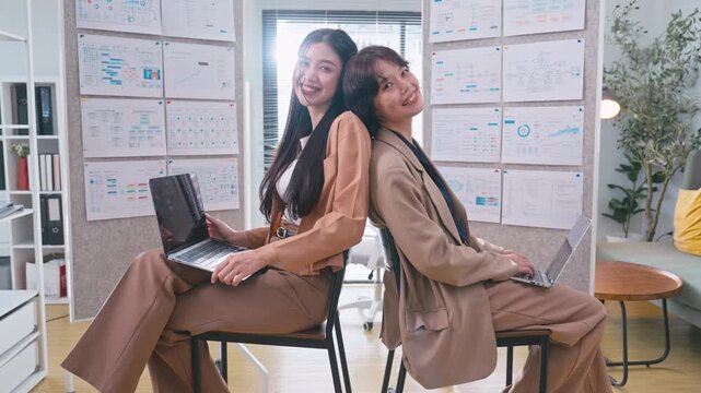 Two confident Asian businesswomen smiling and leaning back to back while sitting on the floor with laptops, showing strong partnership.