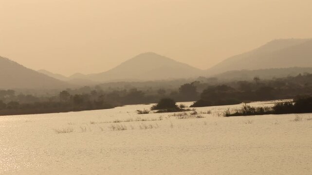 Serene River Landscape with Distant Hills and Silhouetted Trees at Golden Hour Sunset, Calm Water Reflection and Hazy Atmospheric Sky, Peaceful Nature and Wilderness Stock Footage