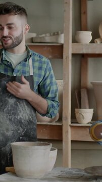 Young man making online video call in pottery workshop while woman making artwork from clay. Communication and creative people concept.