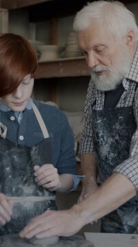 Grandfather teaching curious grandson to work with clay make pots talking and using tools in pottery studio. Professional occupation and family business concept.