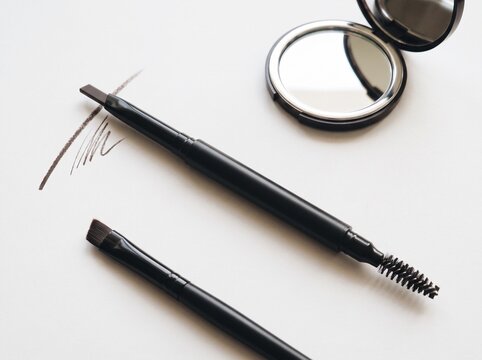 Angled eyebrow brush and a spoolie brush on a white background next to a compact mirror. An eyebrow powder swatch is drawn nearby.