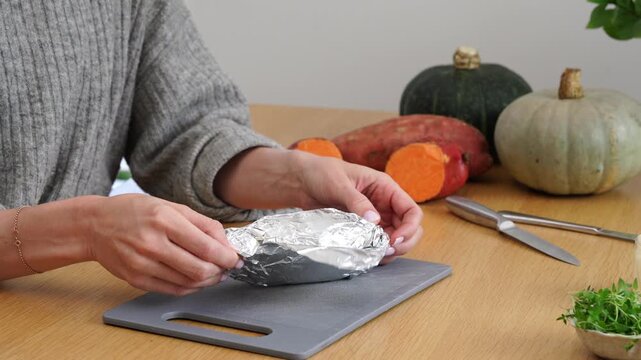 Hands wrap small aluminum foil parcel in autumn kitchen with baked sweet potato on cutting board warm meal prep moment during fall harvest cooking scene closeup wooden table