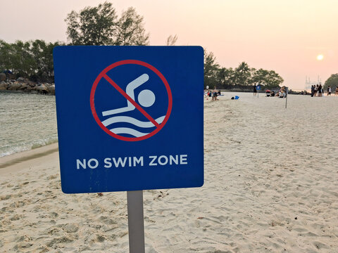 No swim zone sign on a sandy beach at sunset, featuring a swimming prohibition symbol, calm sea, beachgoers, and tropical coastal scenery.