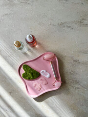 Flat lay of beauty products with jade roller, gua sha, and cosmetic cream textures on a pink plate on marble background. © Jekaterina