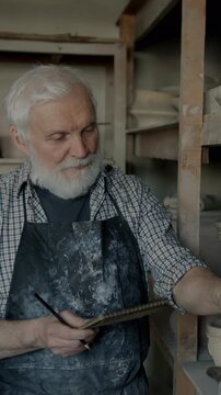 Portrait of senior man pottery business owner checking pots on shelves doing inventory developing studio. People and entrepreneurship concept.