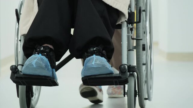 Nurse gently transports female patient with mobility impairment. Doctor pushes wheelchair along clean hospital corridor. Slow motion
