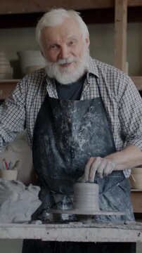 Senior man in apron making pot in workshop and recording video about pottery for internet vlog using smartphone camera. Social media and vlogging concept.