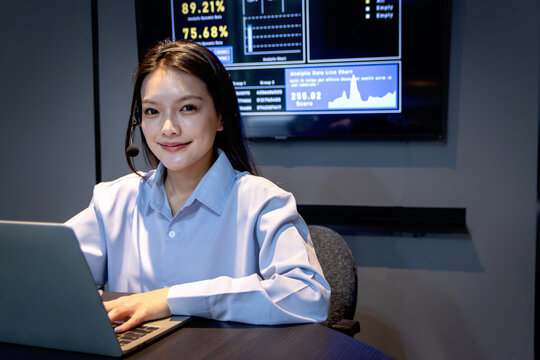 Asian telesale woman with headset works on laptop in late night shift with digital screen background of stock exchange trade marketing information. Happy female telemarketing support customer service.