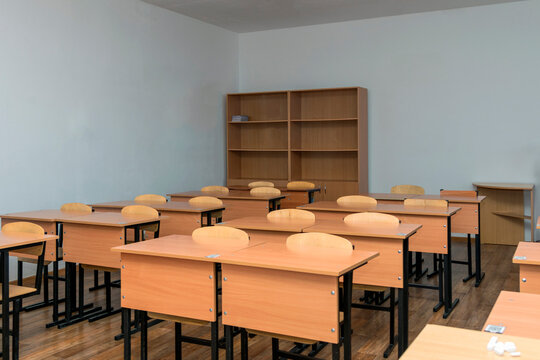 Wide view of an empty school classroom with rows of wooden desks, chairs, and a large bookshelf against a neutral wall