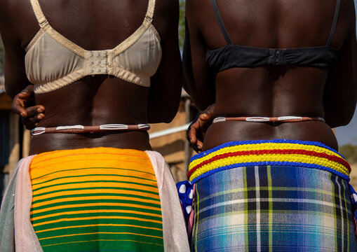 Two women stand side by side from behind wearing lace bras and colorful wrap skirts with beaded belts, Cunene Province, Cahama, Angola