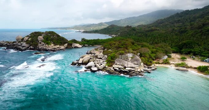 Tayrona National Park, Colombia, revealing a turquoise cove, rocky islets, white surf and tropical shoreline backed by lush jungle landscapes, cinematic aerial view