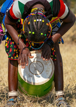 Mudimba tribe woman bending forward to play a drum made from a repurposed metal barrel while wearing a beaded headdress, Cunene Province, Cahama, Angola