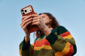 Young woman in striped sweater taking photo with smartphone against blue sky. Close-up of hands...