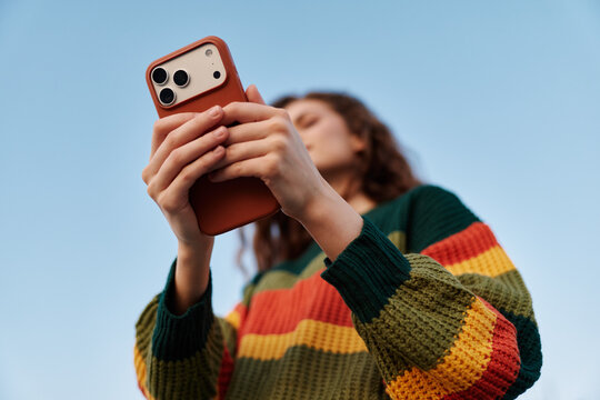 Young woman in striped sweater taking photo with smartphone against blue sky. Close-up of hands holding phone, technology and social media concept.