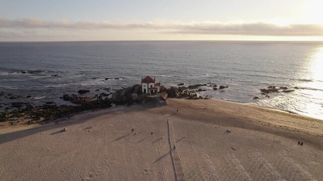 Aerial - Senhor da Pedra chapel sits on granite rocks where Miramar Beach meets the Atlantic in Vila Nova de Gaia Portugal as beachgoers rest on the sand in late afternoon light.