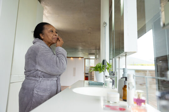 Senior African American woman standing at bathroom counter applying skin cream by mirror