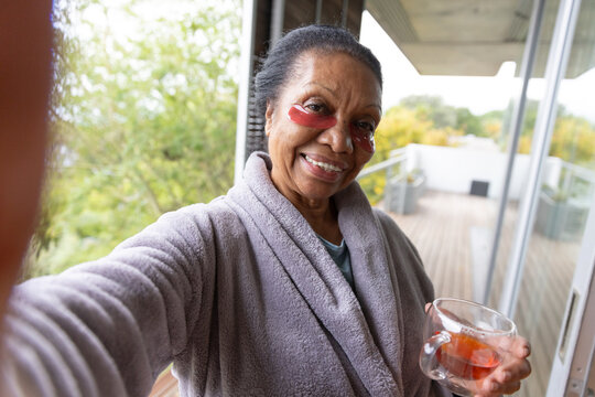 Senior African American woman in gray robe taking selfie on balcony with mug, red under-eye patches