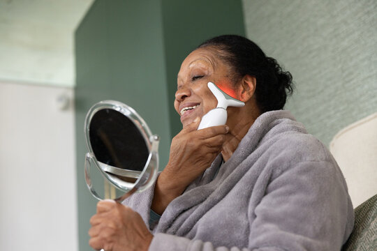 Senior African American woman using mirror, pressing facial device with red LED in bathrobe at home