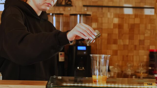 Female barista pours caramel syrup into cup with orange juice at countertop closeup. Barkeeper makes bumblebee cocktail in coffeeshop