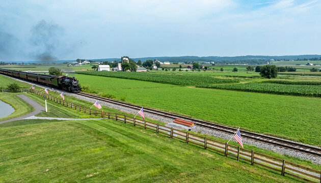 Train Moves Through Green Fields in Rural Area With Flags Lining the Tracks Near Small Farms