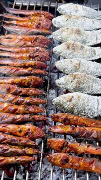 Grilled catfish on skewers and salt-crusted tilapia cooking over hot charcoal at an Asian street food market stall. Fresh barbecue seafood meal prepared outdoors on open fire.