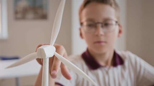 Caucasian boy wearing glasses spinning wind turbine model and taking notes while studying renewable energy. Footage ideal for STEM education campaigns and sustainable technology promotions