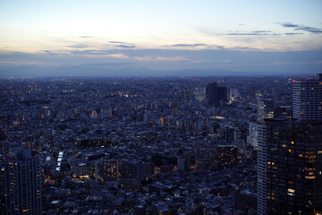 夕暮れ時の高層ビル群が広がる都市の夜景。無数の窓明かりが輝き、遠くには山並みが見える。 © BRUCE